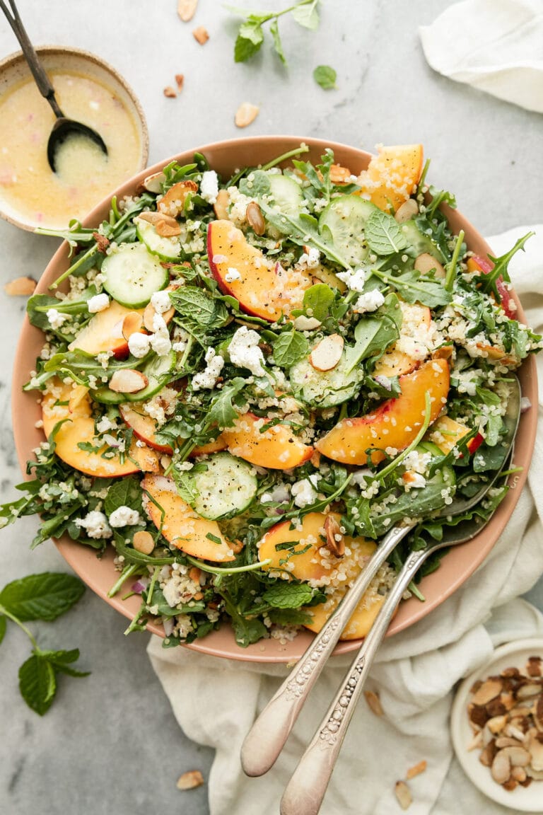 Overhead view of a bowl of quinoa arugula salad with peaches and goat cheese topped with toasted almonds and fresh mint. 