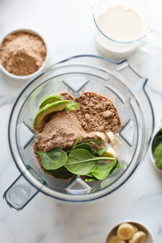 Overhead view of a blender containing chocolate protein powder, avocado, banana slices, and spinach leaves ready for blending. 