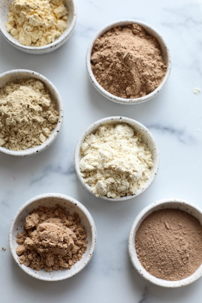 Overhead view of a variety of protein powders in small white bowls. 