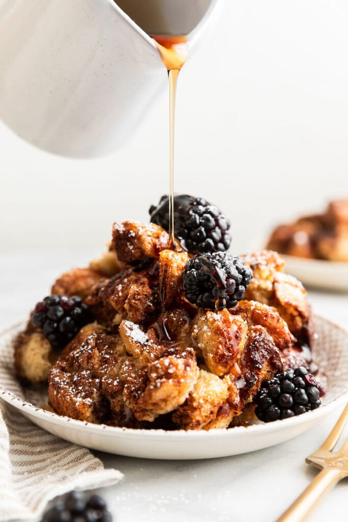 Overhead view of syrup being poured over a plate of French toast topped with powdered sugar and fresh blackberries. 